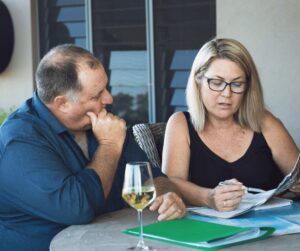 A middle aged couple is sitting at their table looking over maps and booklets, they're also drinking white wine